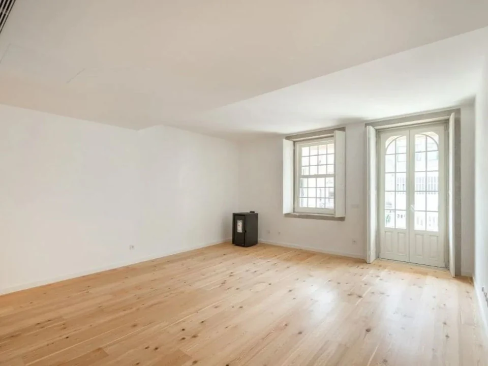 Sala de estar vazia com pavimento de madeira clara, paredes brancas, uma janela com portadas, porta envidraçada e recuperador de calor preto.